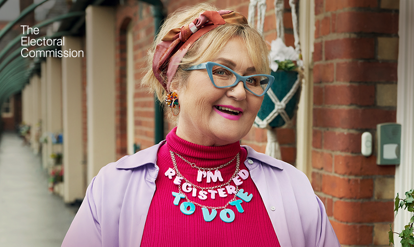 A woman in a bright pink top and lilac jacket with blue glasses with a pink and blue necklace that says 'I'm registered to vote' she is front of a brick building