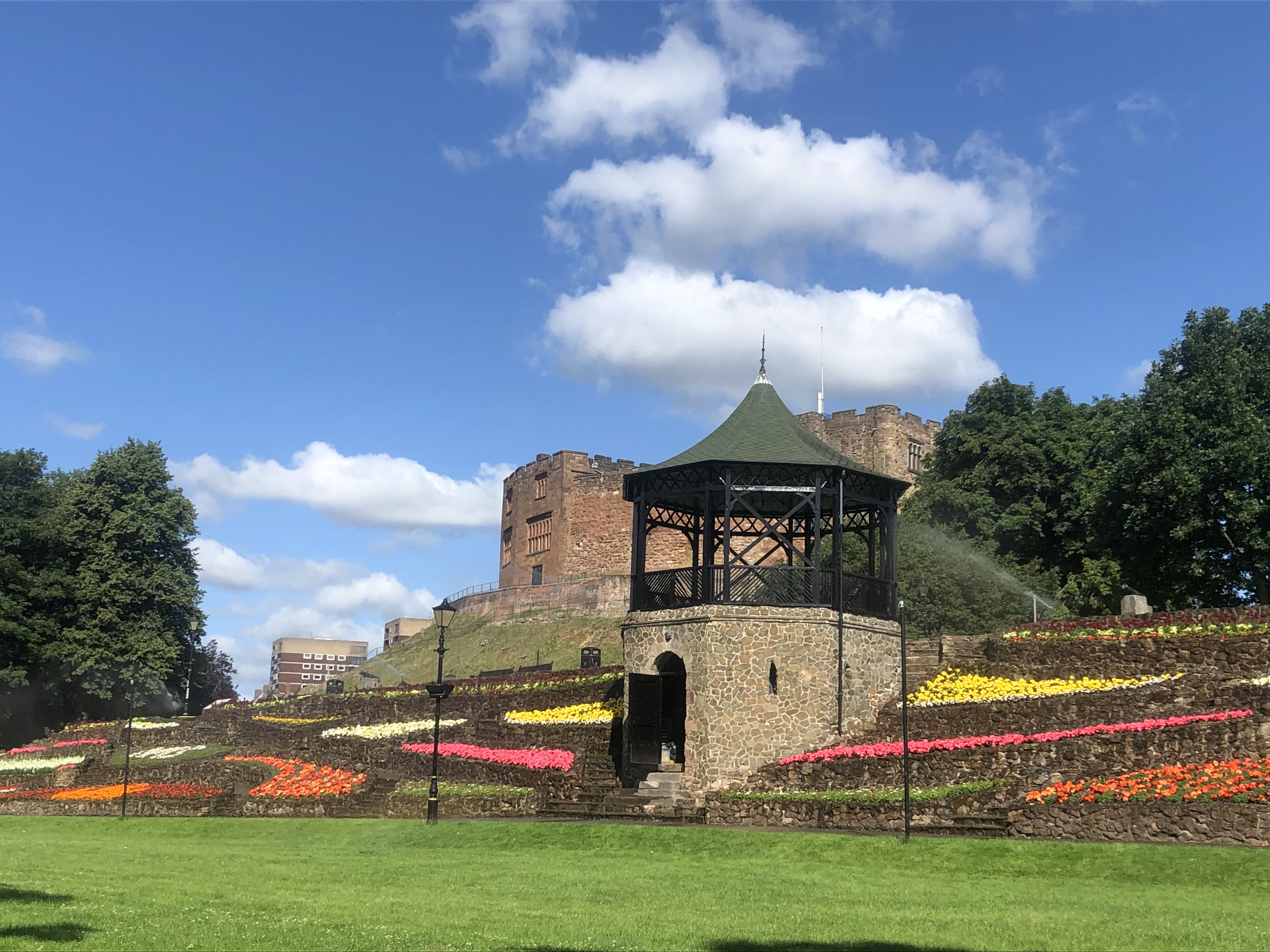 photo of the Castle Grounds with the bandstand and castle in background, blue sky and white clouds plus red, orange and yellow flowers