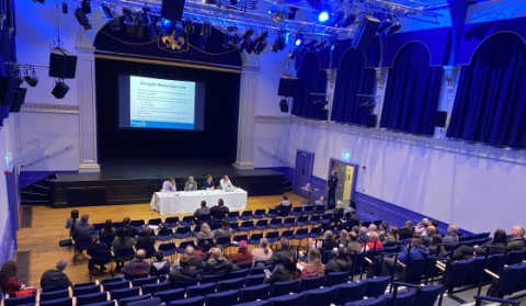 An auditorium with chairs and people sat listening to four people sat at a table at the front